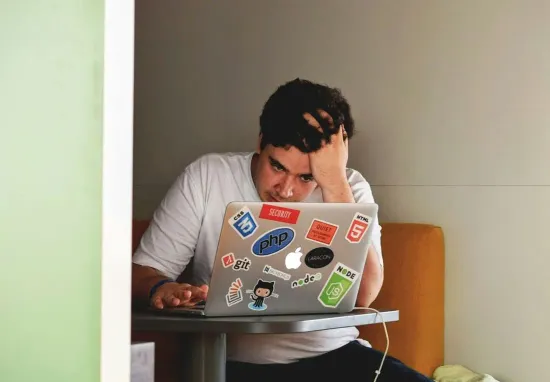 A man with his hand in his hair, frowning at a laptop covered in stickers