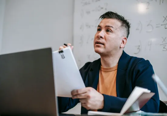 A tutor holding a paper sign-in sheet on a clipboard beside a laptop